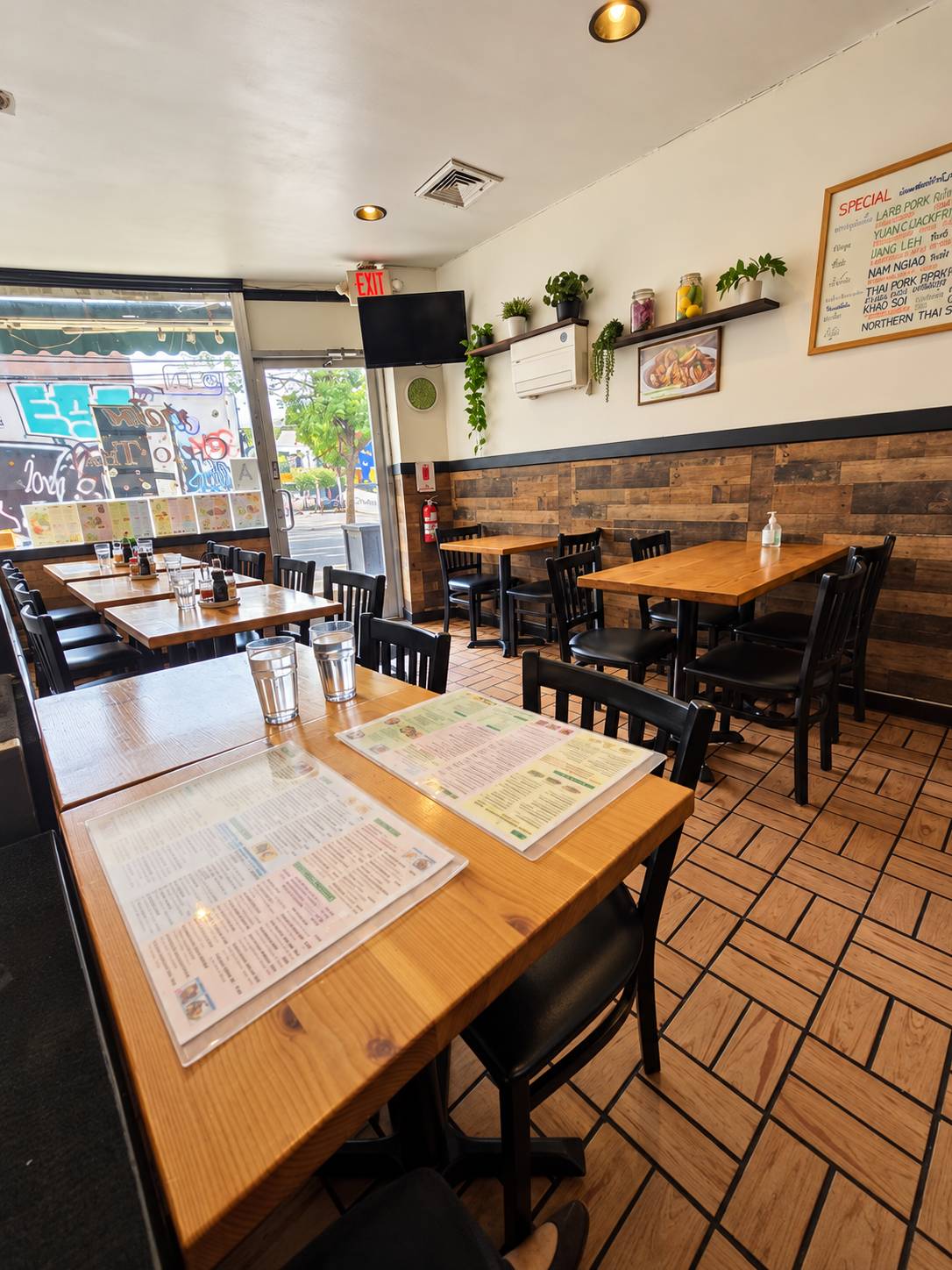Dining room looking toward the glass front door: wood tables with laminated menus, black chairs, plants on a shelf and a specials board.